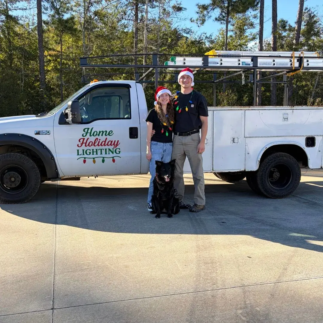 Brandon, Jacqueline, and Aries the dog in Santa hats in front of the Home Holiday Lighting work truck